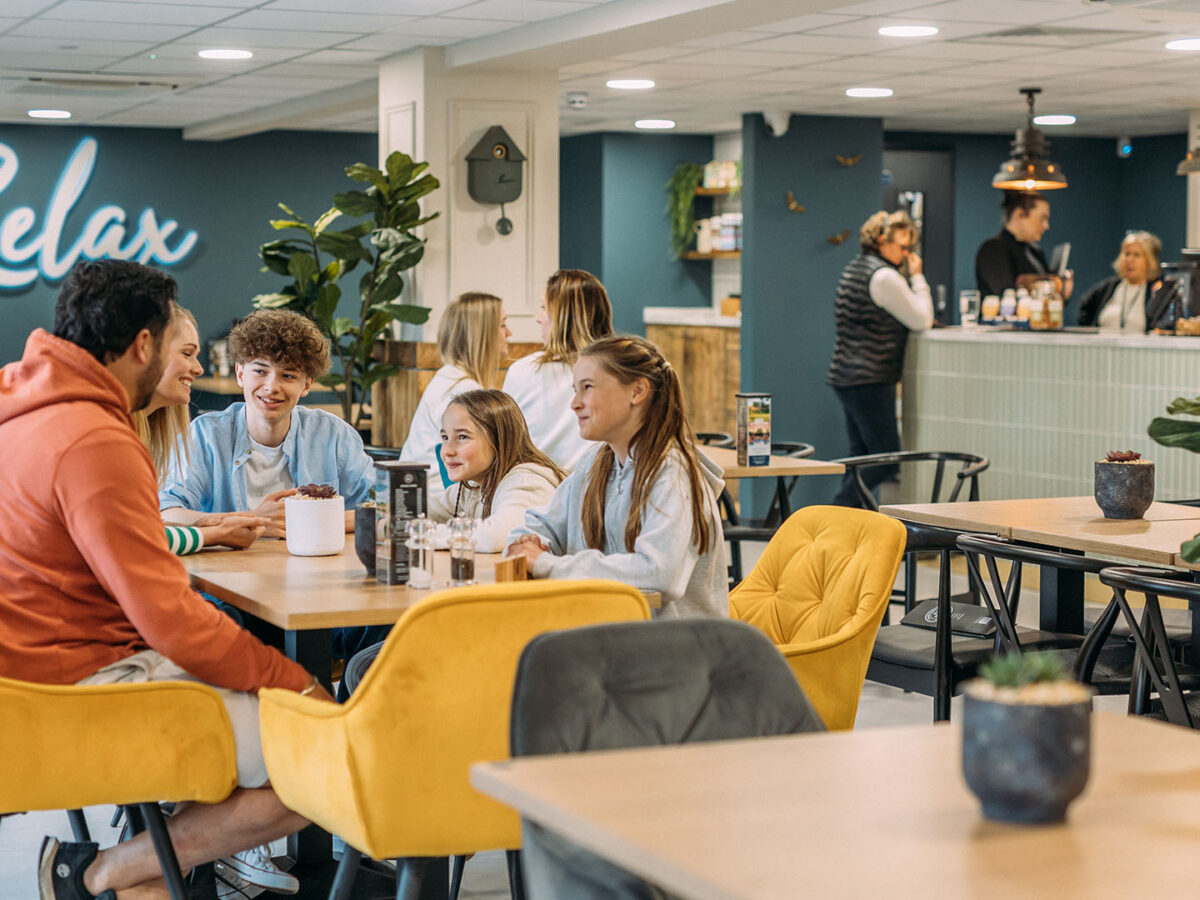 Family seated inside The Hive Cafe, enjoying a meal around a table in a bright and modern setting, with the word Relax in the background on the wall