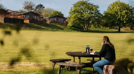 Person enjoying a drink at a picnic table on a grassed field with holiday homes in the background