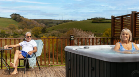 Older woman basking in a hot tub, with older man looking on from a table and chair on an outdoor decking area overlooking the Devon hills