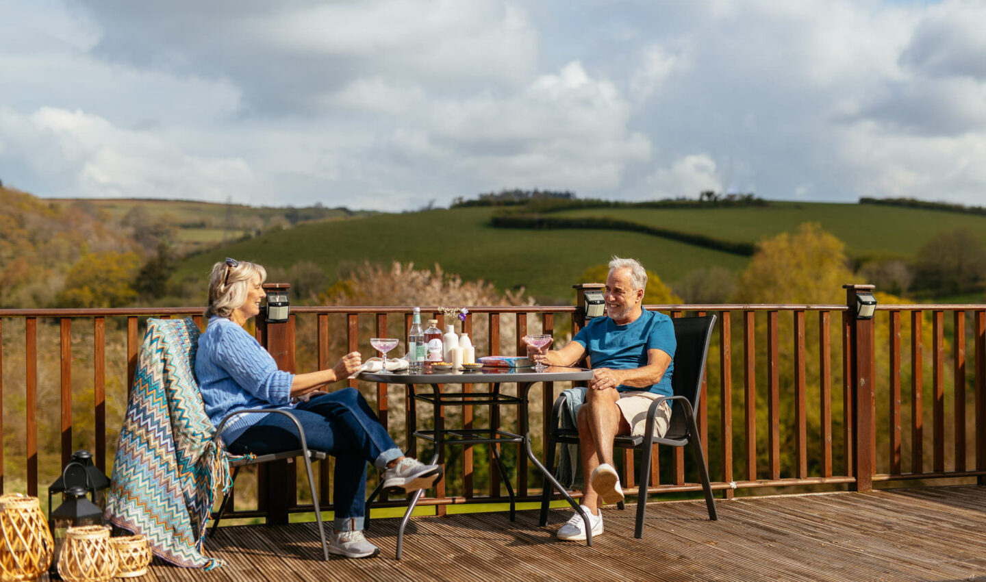 Older couple smiling, sitting at a table sipping on drinks, overlooking the Devon hills, Devon, England.