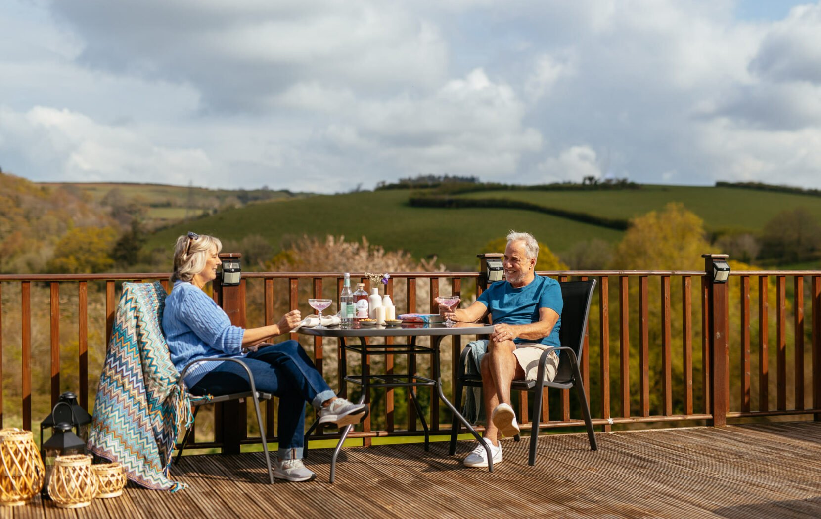 Older couple smiling, sitting at a table sipping on drinks, overlooking the Devon hills, Devon, England.