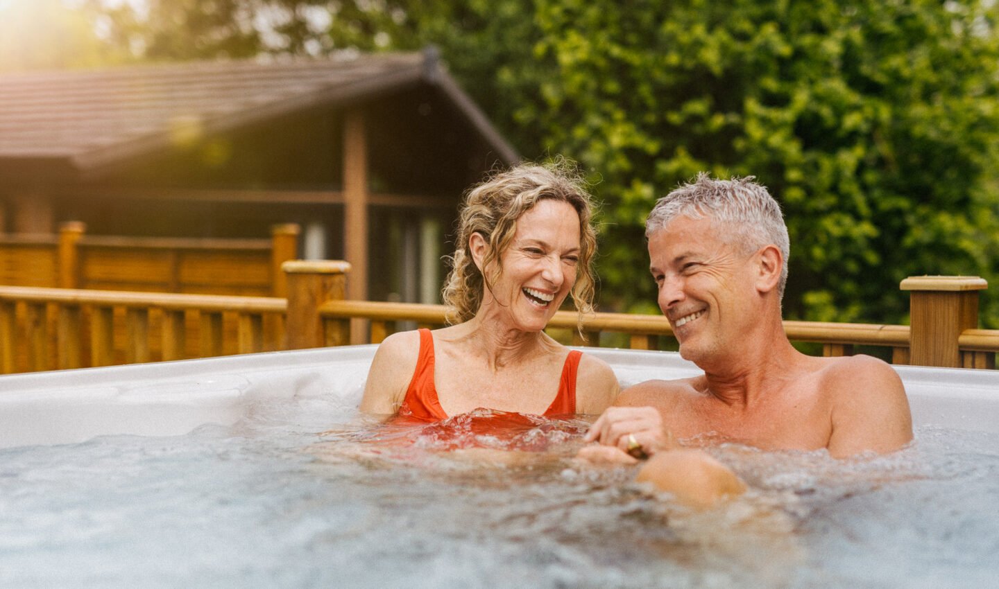 Couple laughing in a steaming bublly hot tub with trees and wooden balcony behind them